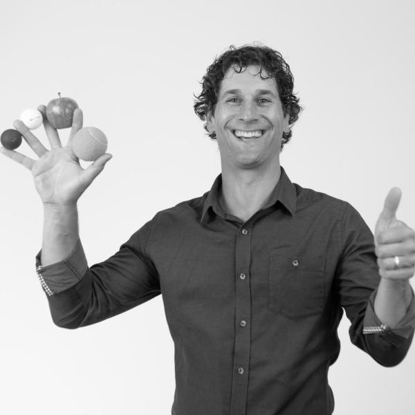 A smiling man holding different size balls and an apple between his fingers. 